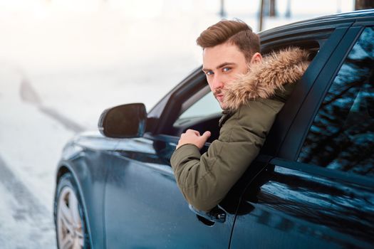 Young adult attractive Caucasian man sits at the wheel of his car sunny winter day. Wintertime road trip. Happy smiling hipster guy sitting in car and looking window. Portrait Positive driver