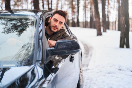 Young adult attractive Caucasian man sits at the wheel of his car sunny winter day. Wintertime road trip. Happy smiling hipster guy sitting in car and looking window. Portrait Positive driver