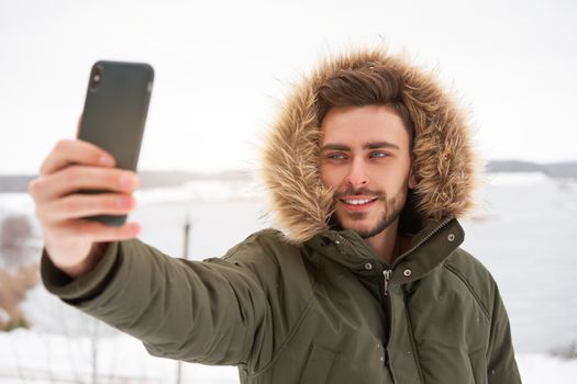 Handsome bearded young caucasian man standing outdoors fur hood winter frozen lake Happy smiling Attractive stylish european guy walking taking selfie on smartphone Photograph Blogger Winter adventure