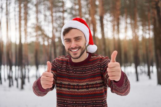 Handsome bearded young caucasian man standing outdoors sweater santa hat winter season forest. Attractive stylish european guy walking snowy christmas woodland Season holiday leisure Showing thumbs up