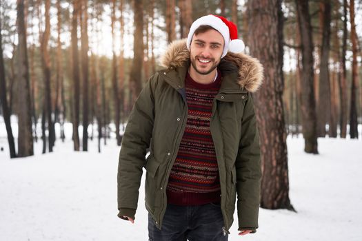 Handsome bearded young caucasian man standing outdoors sweater santa hat winter season forest. Attractive stylish european guy walking snowy christmas woodland Season holiday leisure