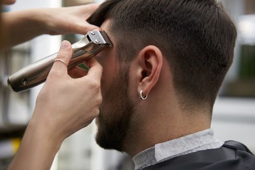 Beautiful caucasian woman hairdresser doing hairstyle to client. Close up portrait female barber with hairdressing tools trimmer at work. hair cutting process Hairstyling in modern barbershop.