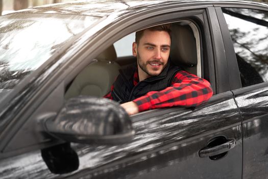 Young adult attractive Caucasian man sits at the wheel of his car sunny winter day. Wintertime road trip. Happy smiling hipster guy sitting in car and looking window. Portrait Positive driver