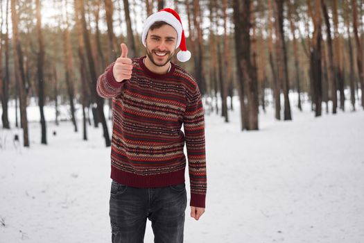 Handsome bearded young caucasian man standing outdoors sweater santa hat winter season forest. Attractive stylish european guy walking snowy christmas woodland Season holiday leisure Showing thumbs up