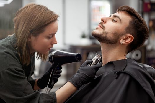 Stylish man sitting barber shop Hairstylist Hairdresser Woman blow dry his hair Portrait handsome happy young bearded caucasian guy getting trendy haircut Attractive barber girl serving client