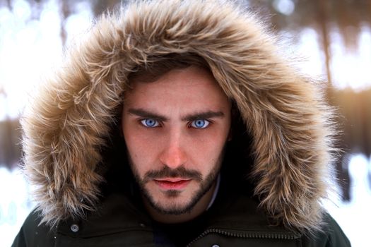 Handsome serious brutal bearded young caucasian man standing outdoors fur hood in winter season forest. Attractive stylish european guy walking snowy christmas woodland Season holiday leisure