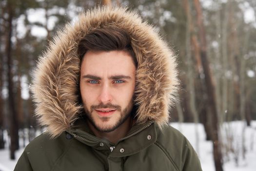 Handsome bearded young caucasian man standing outdoors fur hood in winter season forest. Attractive stylish european guy walking snowy christmas woodland Season holiday leisure