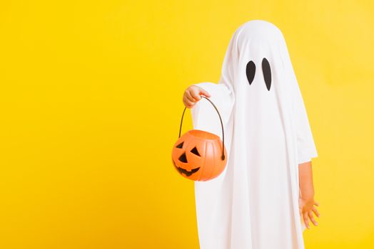 Funny Halloween Kid Concept, little cute child with white dressed costume halloween ghost scary he holding orange pumpkin ghost on hand, studio shot yellow on white background