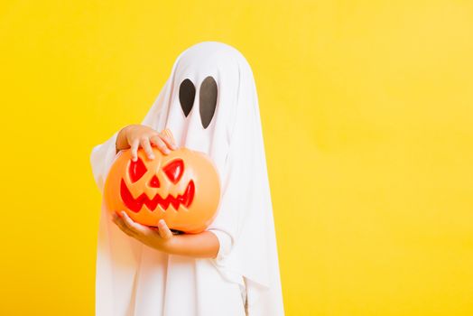 Funny Halloween Kid Concept, little cute child with white dressed costume halloween ghost scary he holding orange pumpkin ghost on hand, studio shot yellow on white background