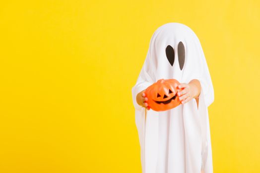 Funny Halloween Kid Concept, little cute child with white dressed costume halloween ghost scary he holding orange pumpkin ghost on hand, studio shot yellow on white background