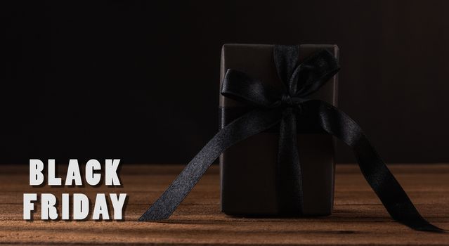 Black Friday sale concept, Gift box wrapped in black paper and black bow ribbon on wooden table studio shot on black background