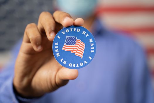 Close up of hands showing I voted by mail sticker with USA flag as background - Concept of US election, mail-in voting or vote by mail.