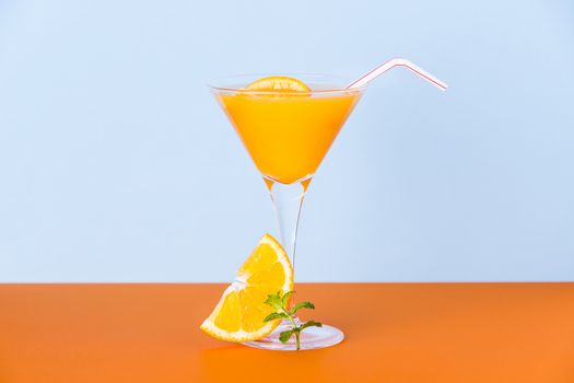 Fresh orange juice in a glass with a slice and a mint leafs at the base on an orange surface and a light blue background