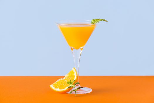 Fresh orange juice with mint leafs and a slice at the base on an orange surface and a light blue background