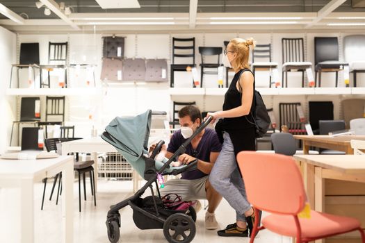Young family with newborn in stroller shopping at retail furniture and home accessories store wearing protective medical face mask to prevent spreading of corona virus when shops reopen.