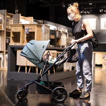 Young mom with newborn in stroller shopping at retail furniture and home accessories store wearing protective medical face mask to prevent spreading of corona virus. New normal during covid epidemic.