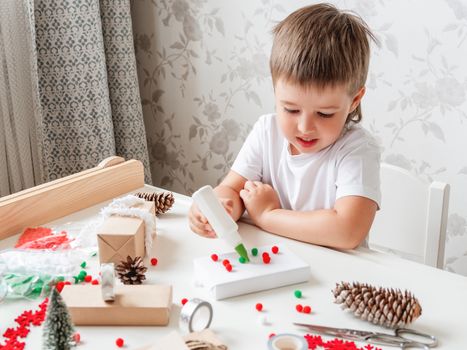Child wrapped Christmas presents in craft paper with white fluffy pompons, feathers and fringe as decoration. White table with handmade New Year gifts.