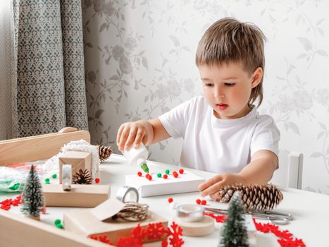 Child wrapped Christmas presents in craft paper with white fluffy pompons, feathers and fringe as decoration. White table with handmade New Year gifts.