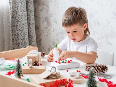Child wrapped Christmas presents in craft paper with white fluffy pompons, feathers and fringe as decoration. White table with handmade New Year gifts.