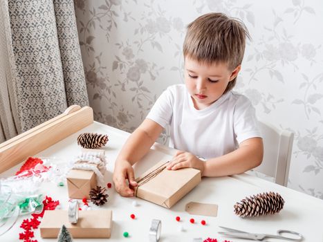 Kid wraps handmade Christmas presents in craft paper with colorful pompons and snowflake ribbons. Child prepares gifts for New Year celebration. Peaceful leisure activity before winter holidays.