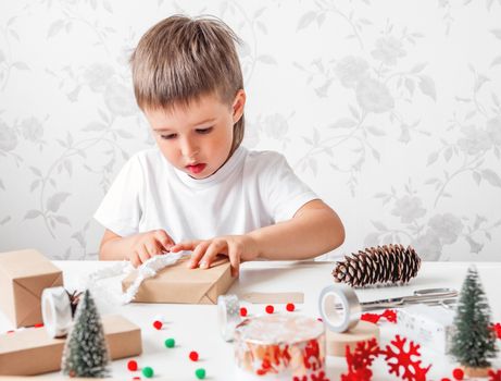 Kid wraps handmade Christmas presents in craft paper with colorful pompons and snowflake ribbons. Child prepares gifts for New Year celebration. Peaceful leisure activity before winter holidays.
