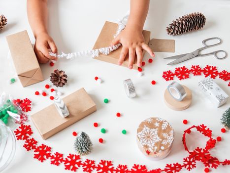 Kid wraps handmade Christmas presents in craft paper with colorful pompons and snowflake ribbons. Child prepares gifts for New Year celebration. Peaceful leisure activity before winter holidays.