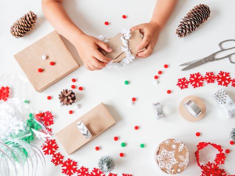 Kid wraps handmade Christmas presents in craft paper with colorful pompons and snowflake ribbons. Child prepares gifts for New Year celebration. Peaceful leisure activity before winter holidays.