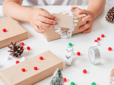 Kid wraps handmade Christmas presents in craft paper with colorful pompons and snowflake ribbons. Child prepares gifts for New Year celebration. Peaceful leisure activity before winter holidays.