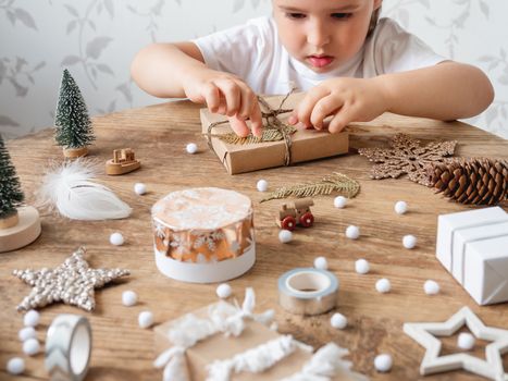 Child wrapped Christmas presents in craft paper with white fluffy pompons, feathers and fringe as decoration. Wooden table with hand made New Year gifts.