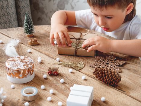 Child wrapped Christmas presents in craft paper with white fluffy pompons, feathers and fringe as decoration. Wooden table with hand made New Year gifts.