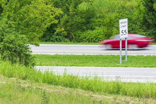 Horizontal shot of a speeding red car streaking by a speed limit sign.  Blurring shows motion.