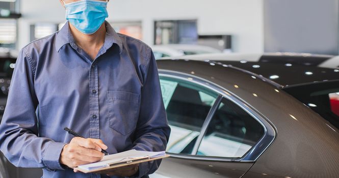 Asian saleman wear surgical mask working in inspector checking writing on clipboard in garage of dealership Wait mechanic In showroom car and insurance new normol covid-19 in showroom dealership