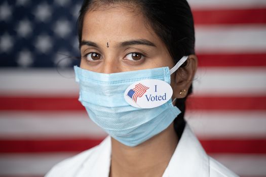 Girl with I voted sticker on medical face mask with US flag as background - concept of US election