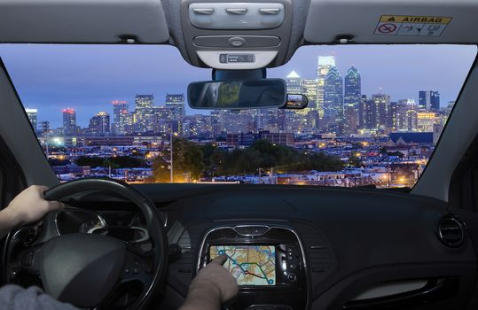 Driving a car while using the touch screen of a GPS navigation system towards Philadelphia skyline at night as seen from the Stadium District, USA
