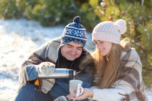 Young couple in love drink a hot drink from a thermos, sitting in the winter in the forest, tucked into warm, comfortable rugs, and enjoy nature. The guy pours a drink from a thermos in a cup.
