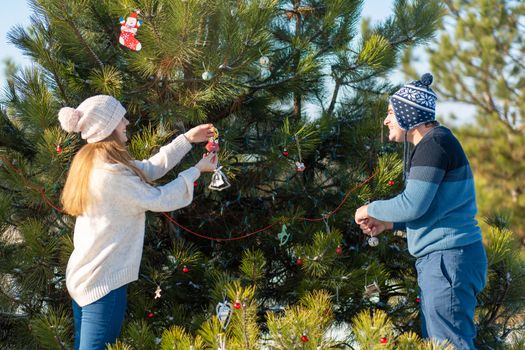 a guy with a girl decorates a green Christmas tree on a street in the winter in the forest with decorative toys and garlands. Christmas tree decorations.