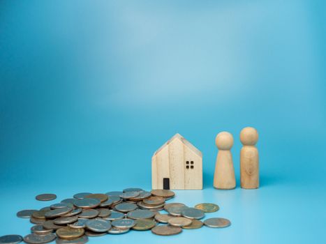 A wooden doll standing next to a replica wooden house and a pile of money has a blue background. Home buying and selling ideas.