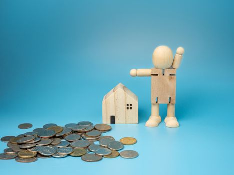 A wooden doll standing next to a replica wooden house and a pile of money has a blue background. Home buying and selling ideas.