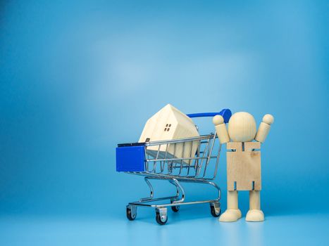 Wooden dolls that Stand next to the shopping cart With a model wooden house on top And has a blue background. Home buying and selling ideas