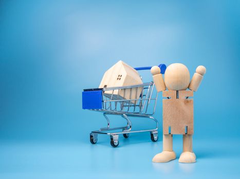 Wooden dolls that Stand next to the shopping cart With a model wooden house on top And has a blue background. Home buying and selling ideas