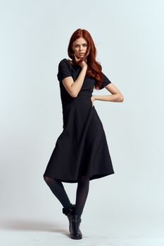 A woman in a black dress on a light background and pantyhose shoes red hair and pose in full growth. High quality photo