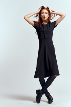 A woman in a black dress on a light background and pantyhose shoes red hair and pose in full growth. High quality photo