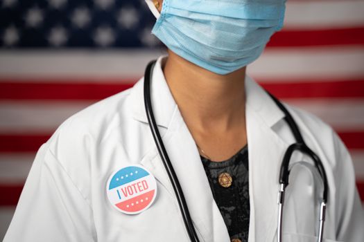 Doctor or nurse medical mask and stethoscope wearing placing I voted sticker to her apron - concept of US election with US flag as background
