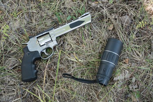 A telescope or binoculars are lying in the forest on the grass next to a pistol revolver.