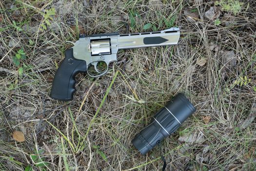 A telescope or binoculars are lying in the forest on the grass next to a pistol revolver.