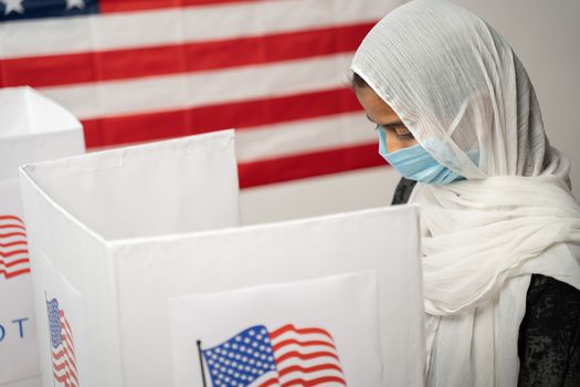 Girl with Hijab or head covering and mask worn busy at polling booth with US flag as background - Concept of US election