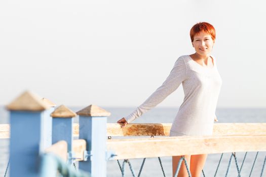 Wide smiling young woman with red short hair cut stays on wooden pier. Coastal morning sunlight. Natural beauty.