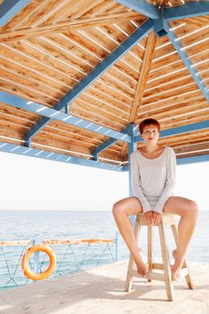 Young woman with red short hair cut sits on wooden pier at lifeguard's place. Coastal morning. Natural beauty.