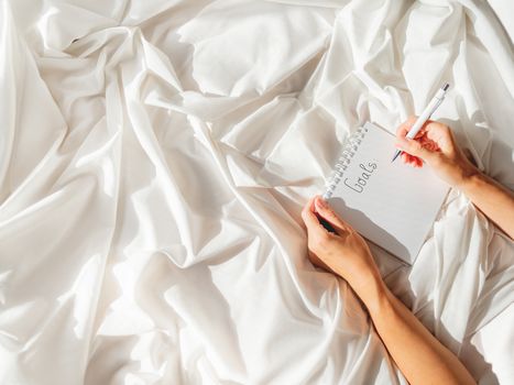 Woman is preparing to write list of her goals. Top view on crumpled bed sheet and notepad with white paper pages. Practice of positive writing at morning after waking up. Flat lay with copy space.