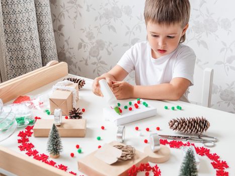 Kid wraps handmade Christmas presents in craft paper with colorful pompons and snowflake ribbons. Child prepares gifts for New Year celebration. Peaceful leisure activity before winter holidays.